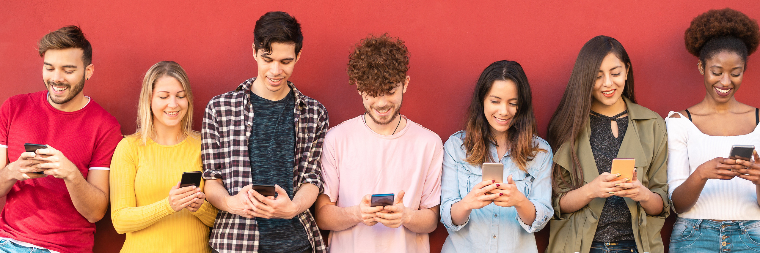 Eine Gruppe junger Leute, nebeneinander an einer roten Mauer lehnend, mit Handys in der Hand 