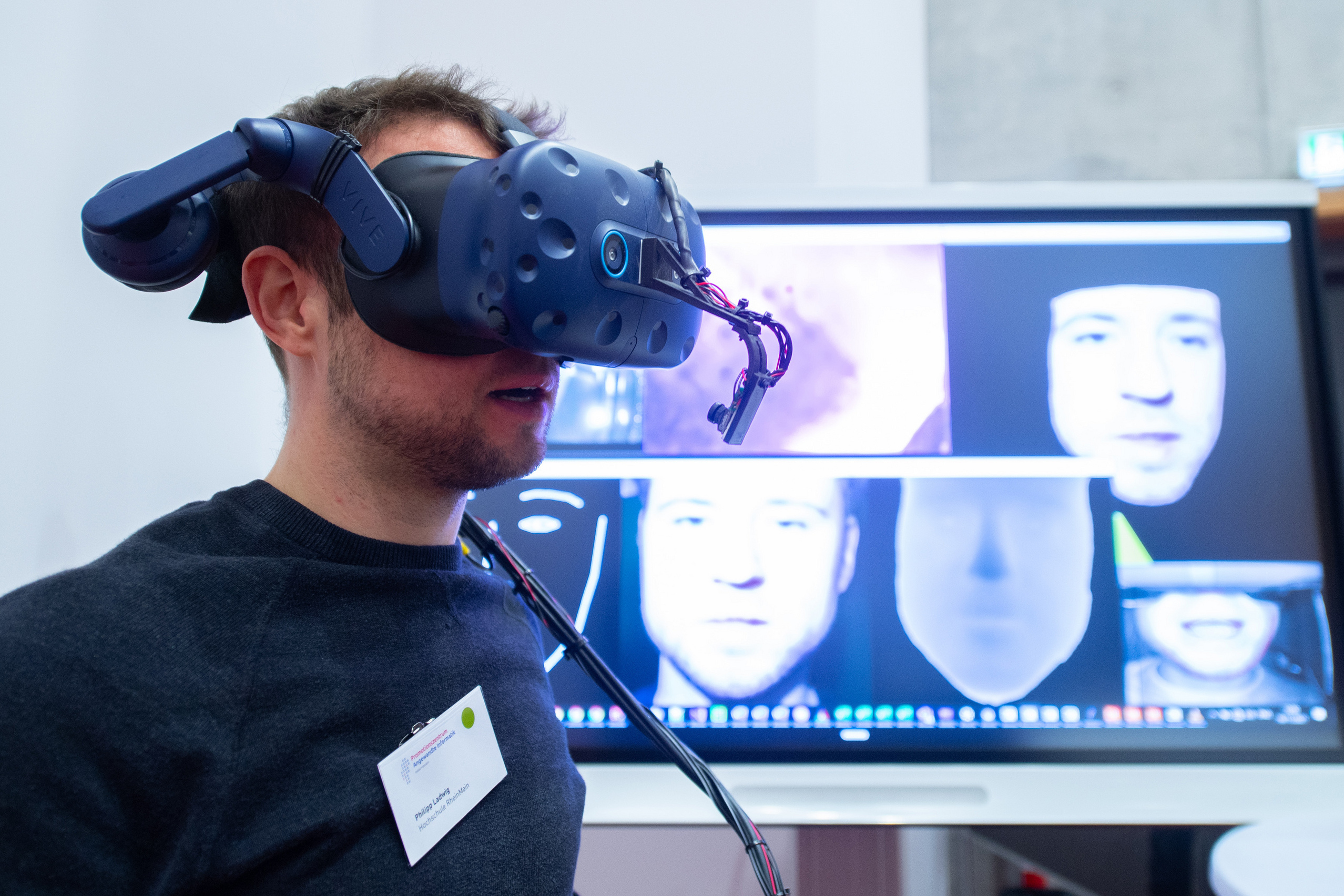 Junger Mann mit Virtual-Reality-Brille im Gesicht, im Hintergrund ein großer Bildschirm. 