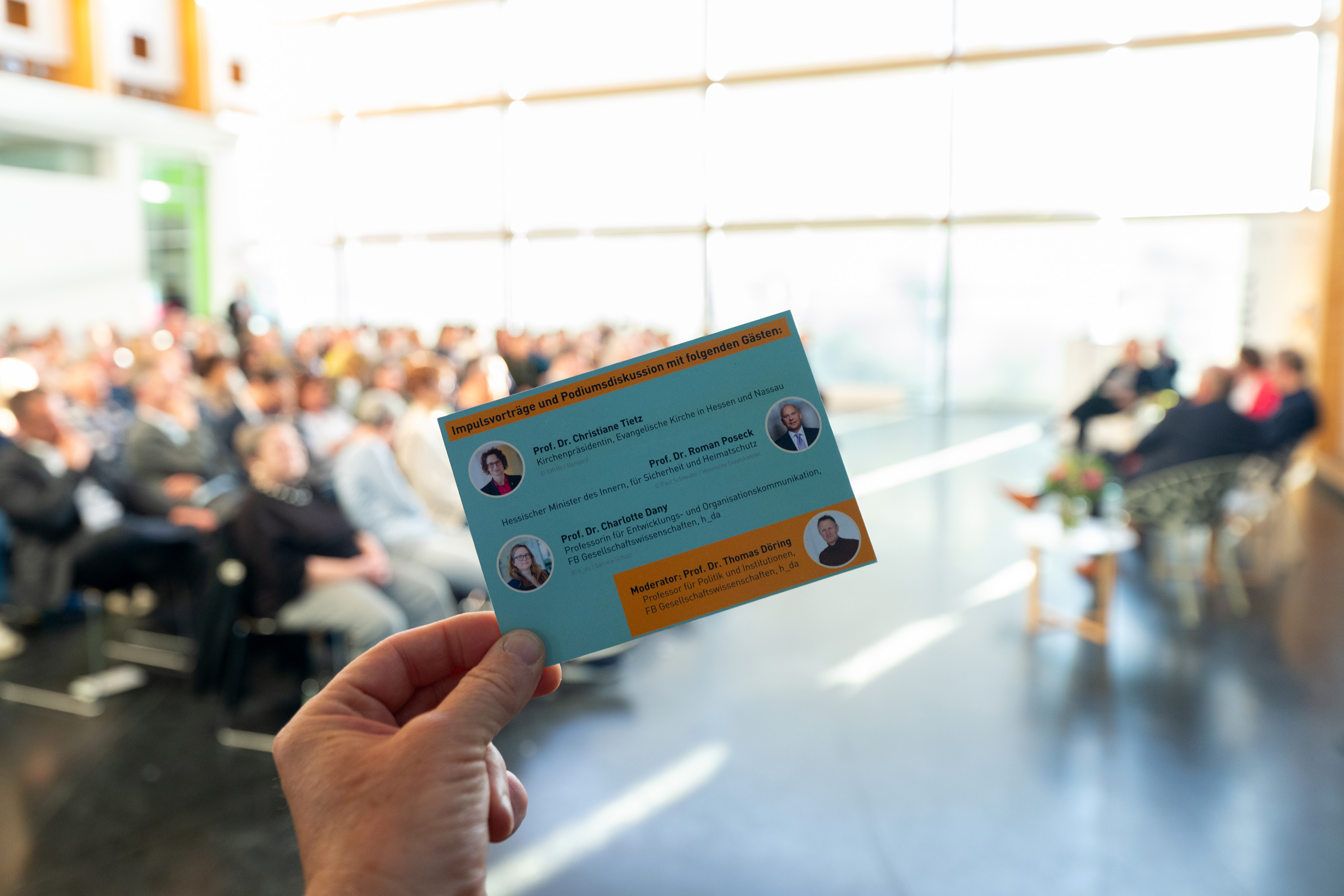 Die Einladungskarte zum Dialog-Forum im Vordergrund, dahinter unscharf das Auditorium.
