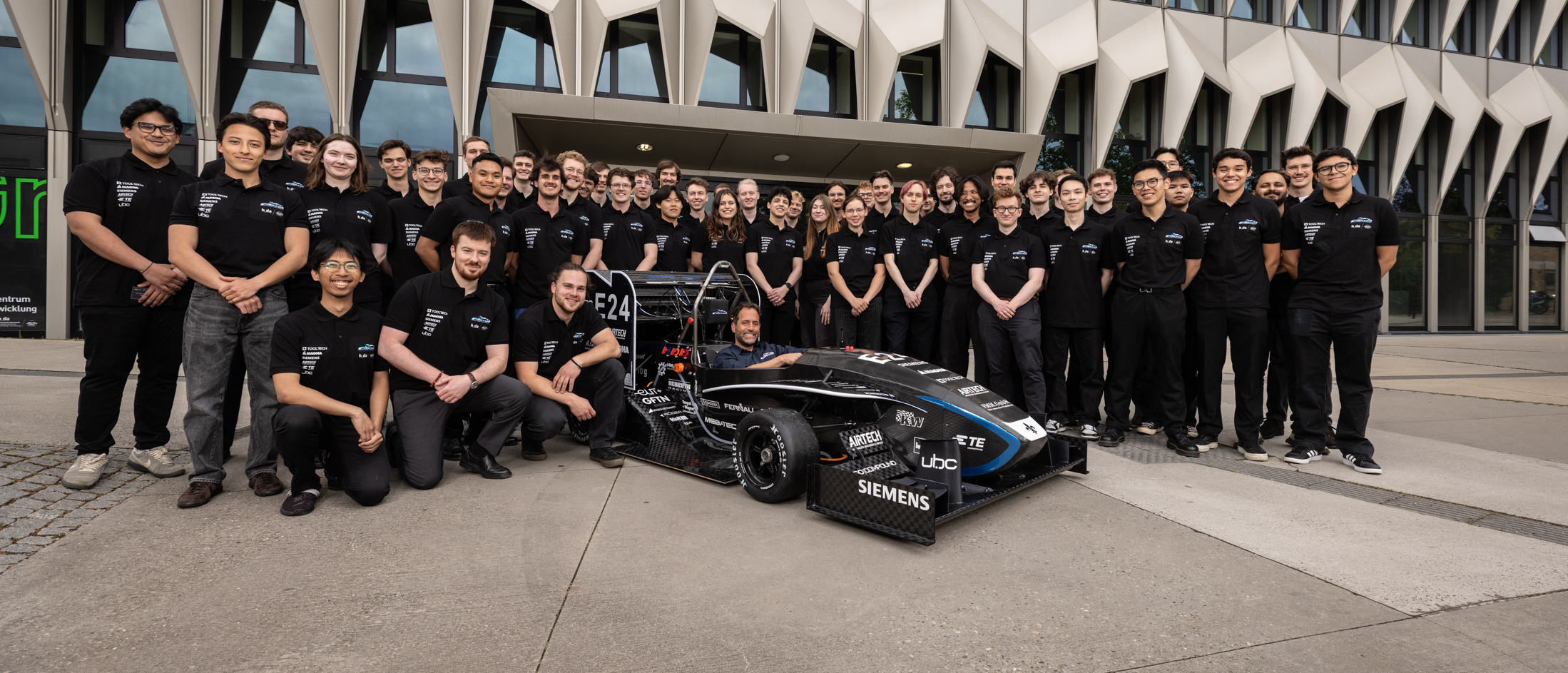 Gruppenfoto mit allen Team-Mitgliedern in Schwarz gekleidet hinter dem Rennwagen stehend, vor dem h_da-Hochhaus