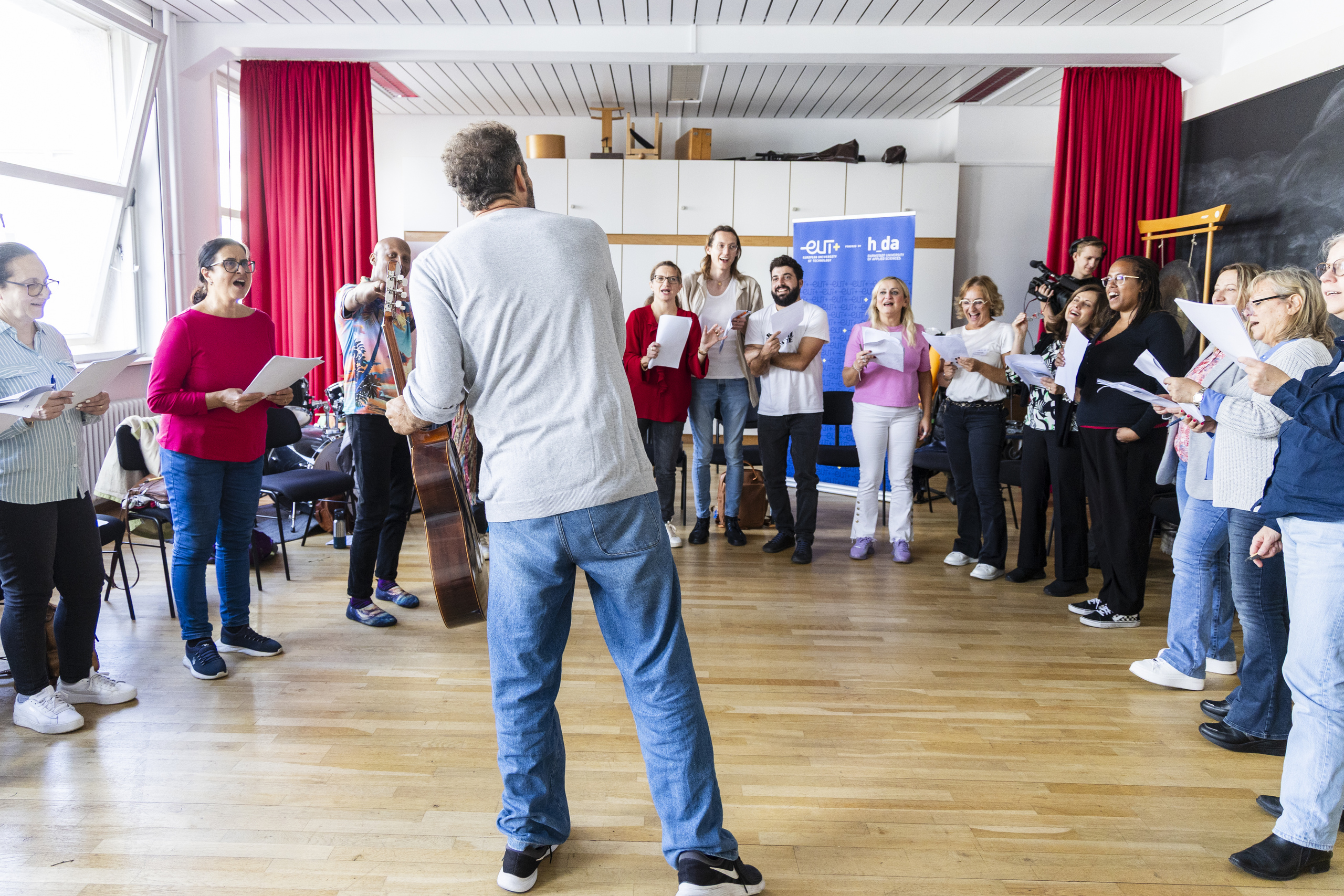 Yiannis Christidis mit Gitarre von hinten vor der im Halbkreis stehenden Chorgruppe