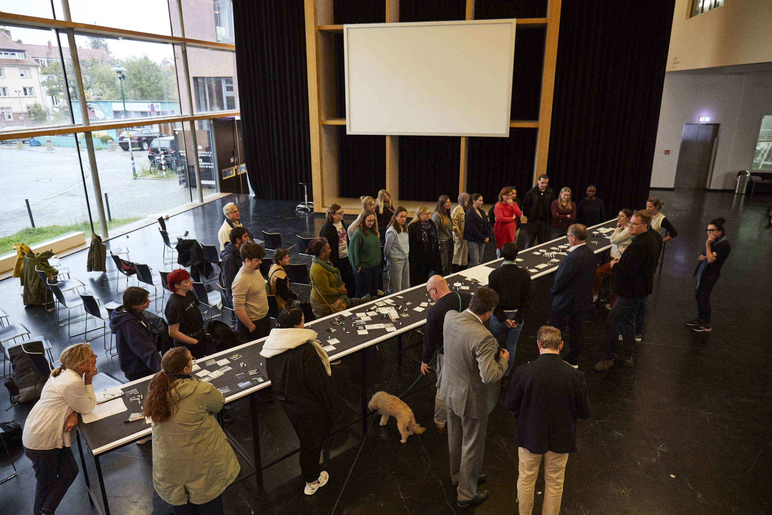 Workshop-Teilnehmende stehen um einen sehr lang gezogenen Tisch herum und interagieren in einer Workshopsituation miteinander