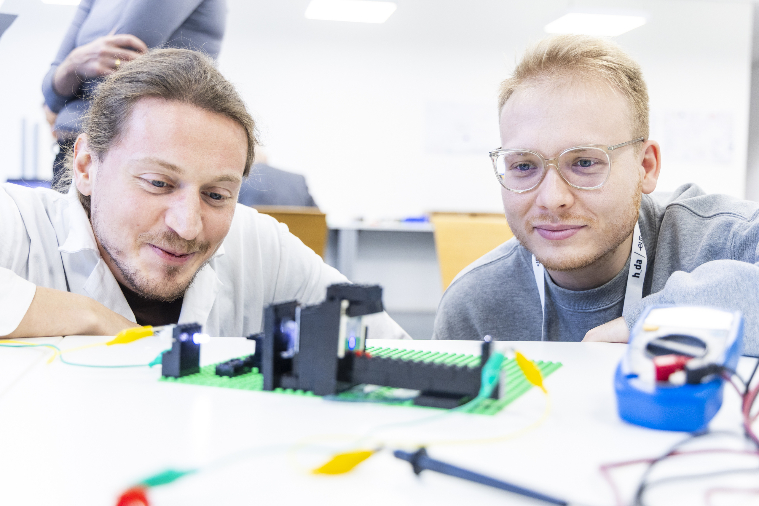 h_da-Mitarbeiter Moritz Schattmann kniet mit einem Workshopteilnehmer vor einem Tisch mit einem Lego-Experiment. 