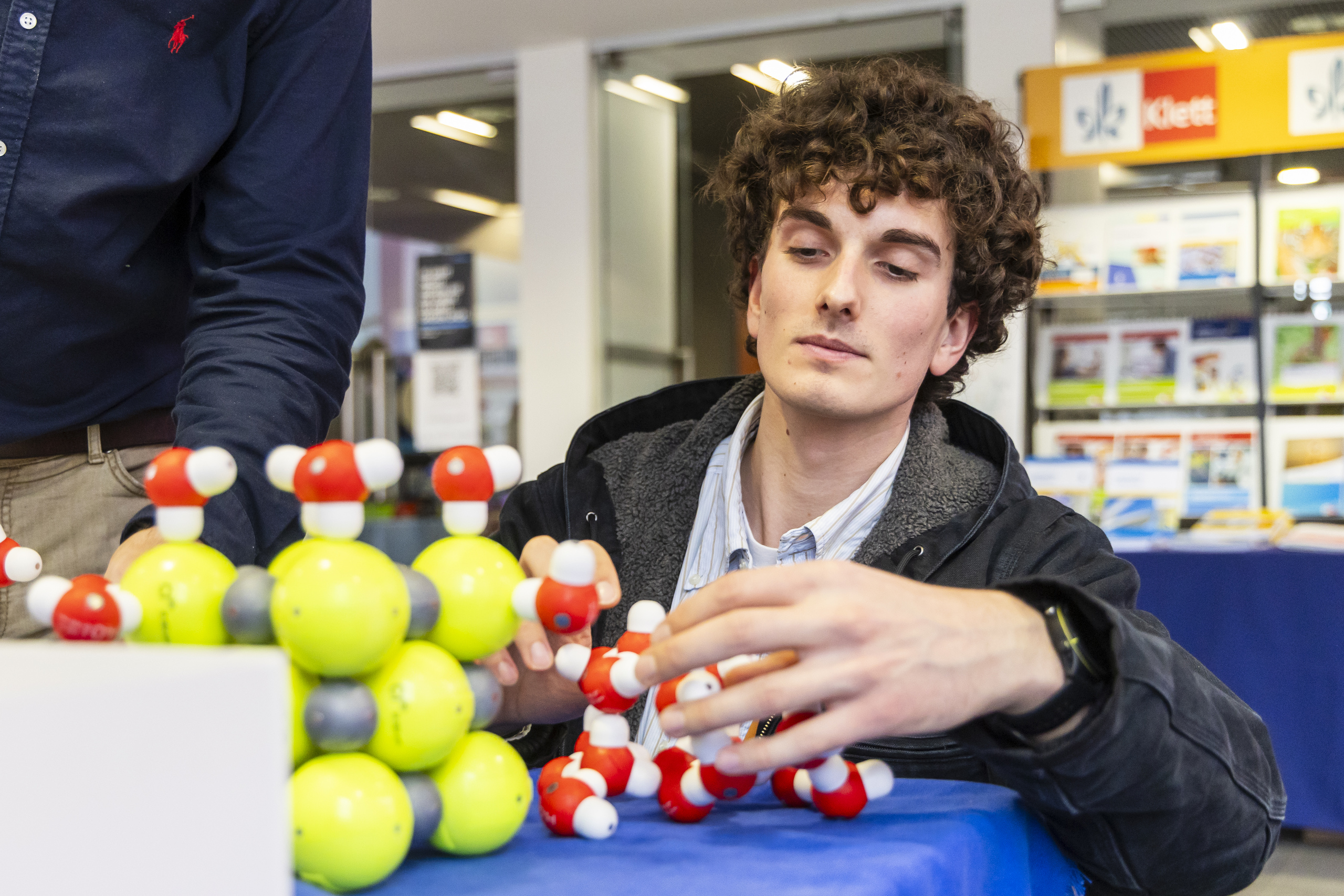 Junger Mann kniet vor einem Tisch mit Molekülmodellen aus bunten Kugeln