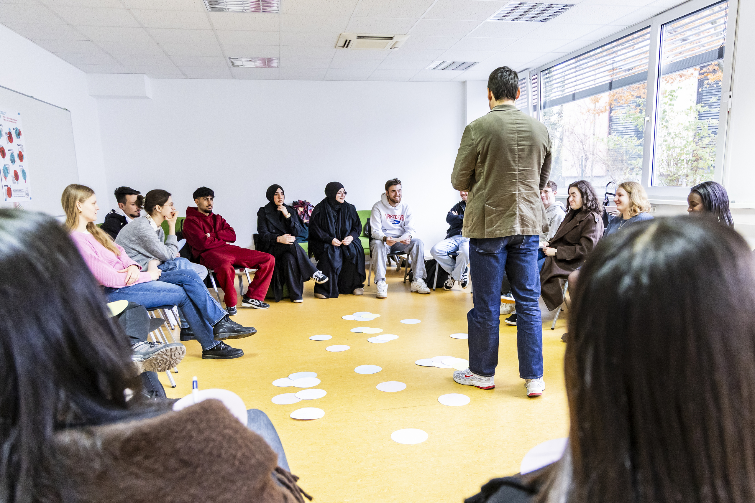 Studierende sitzen im Stuhlkreis, TRainer in der Mitte, auf dem Boden liegen beschriftete Pappschilder