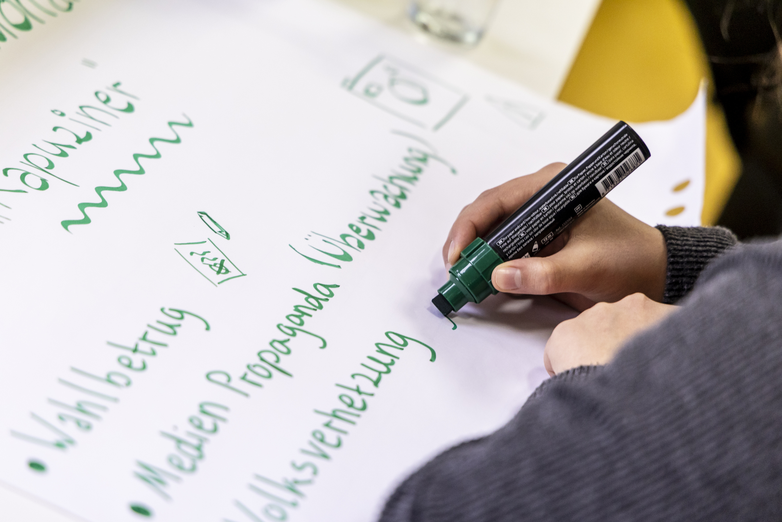 Blick über die Schulter einer teilnehmenden Person mit Stift in der Hand, die auf ein Blatt Papier schreibt