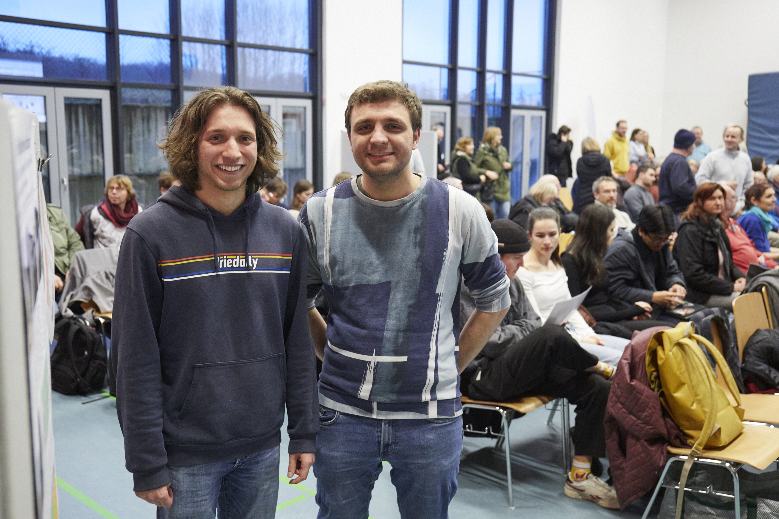 Zwei Studierende stehen nebeneinander und lächeln in die Kamera, im Hintergrund Stuhlreihen mit vielen Menschen