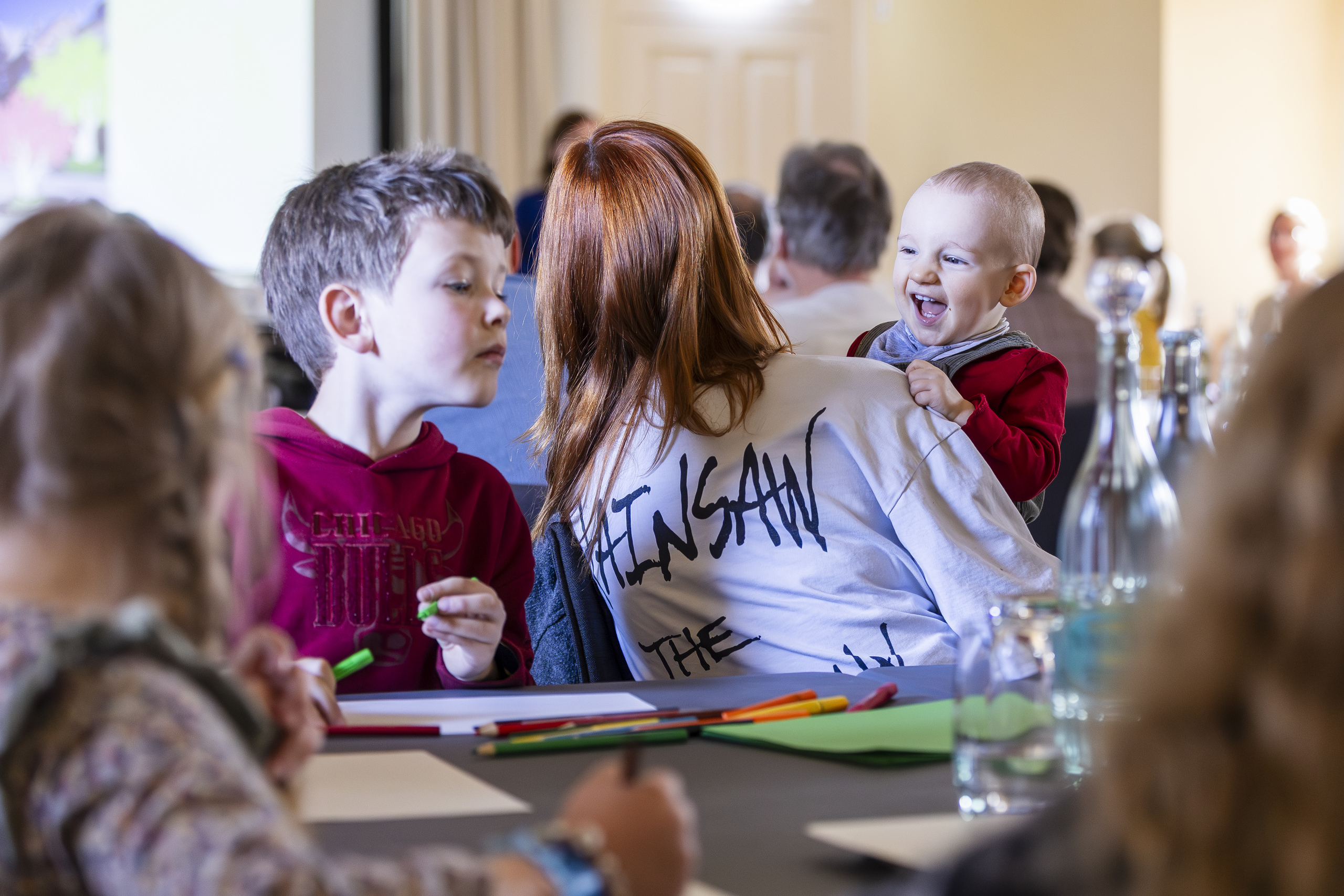 Junge Frau von hinten mit Baby auf dem Schoß, darum herum Kinder an Tischen im Gemeindesaal
